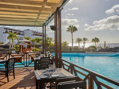 Terrazza dell'hotel con tavoli accanto alla piscina e vista su palme e mare al tramonto.