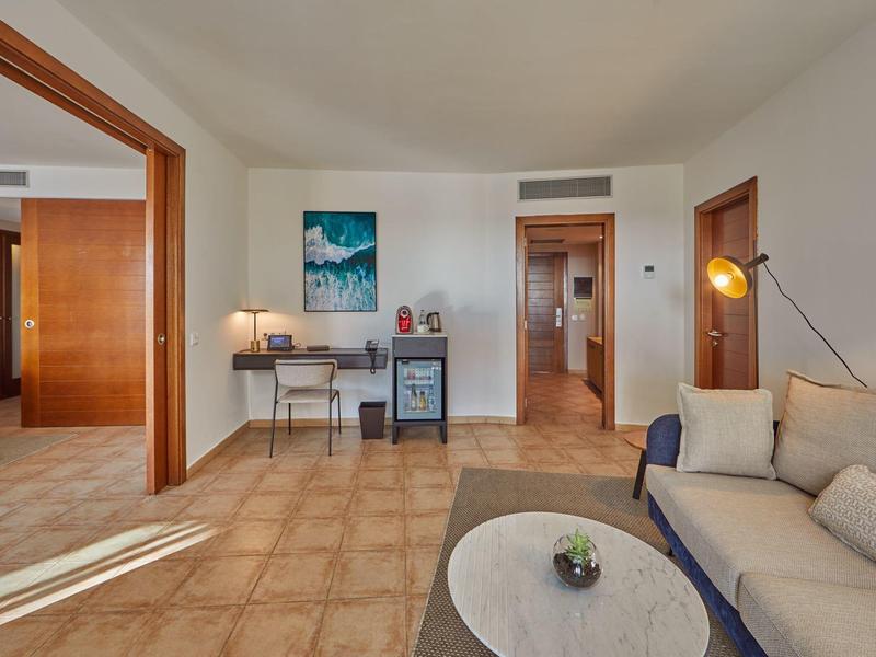 Area accogliente della camera d'hotel con divano, tavolo, scrivania e minibar accanto a un corridoio.