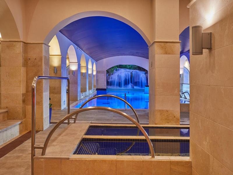 Area piscina interna con vasche idromassaggio riscaldate e grande piscina sotto una volta blu.