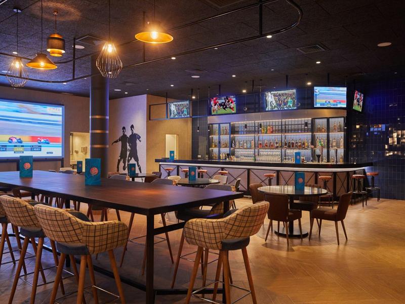 Bar d'hotel moderna con sedie eleganti, tavoli, scaffali illuminati e grandi schermi.