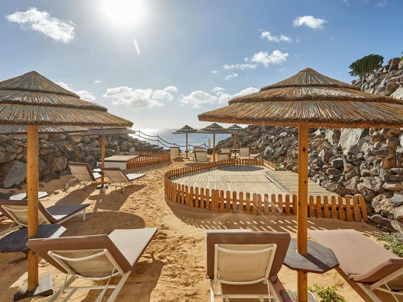 Area spiaggia soleggiata con ombrelloni di paglia e lettini, circondata da rocce.