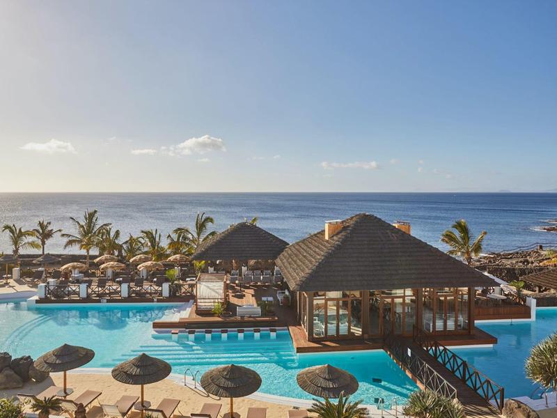 Resort con grande piscina, palme e bar sulla spiaggia sotto cielo blu vicino al mare.