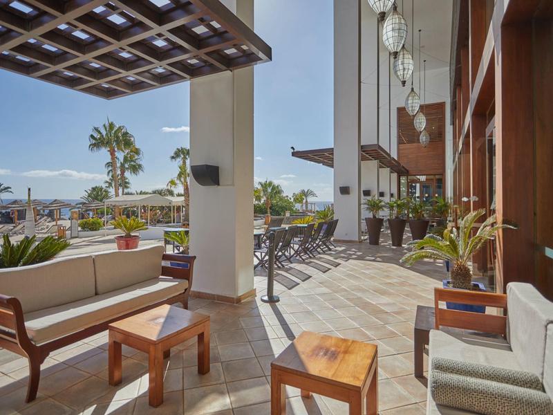 Terrazza coperta dell'hotel con mobili in legno, palme e vista sulla piscina e montagna.