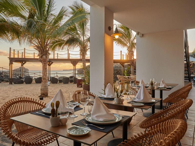 Ristorante sulla spiaggia con tavoli apparecchiati e vista su palme e mare al tramonto.