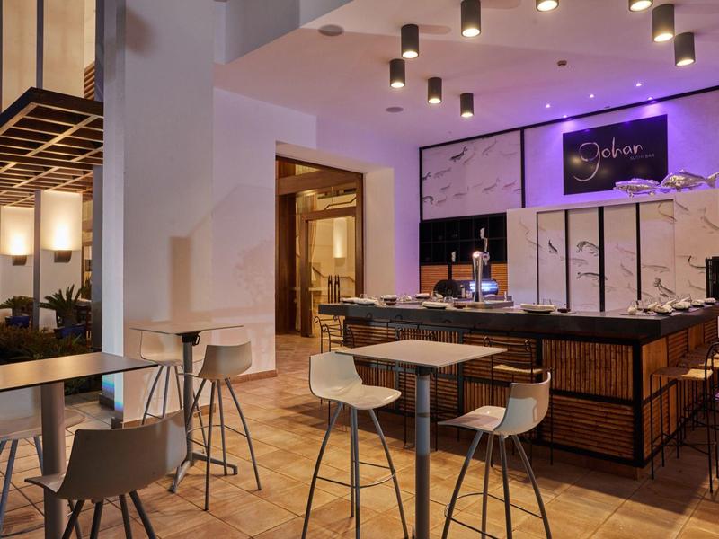 Area bar moderna in hotel con bancone in legno, sgabelli e tavoli, illuminata da luci a soffitto.