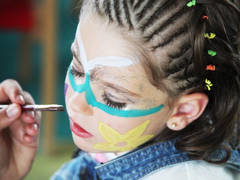 Kind krijgt kleurrijke schmink op het gezicht, draagt een spijkerjasje en heeft gevlochten haar met kralen.