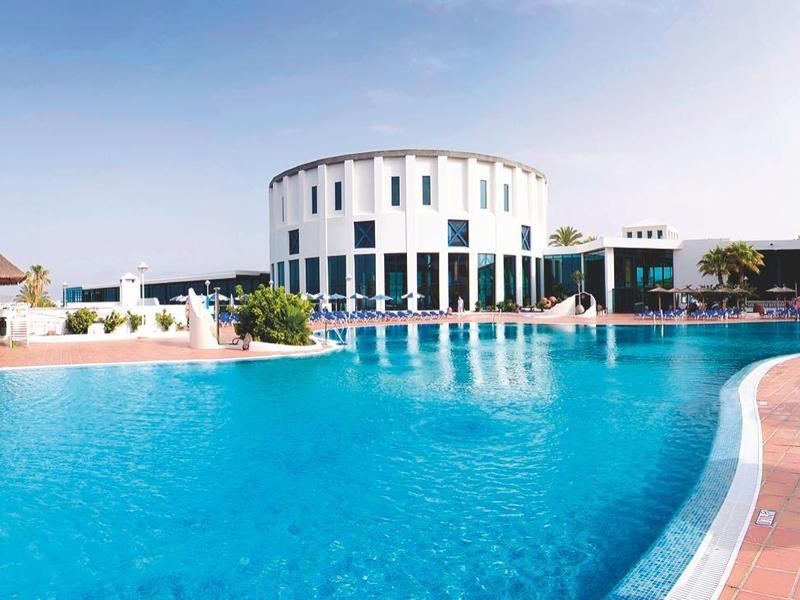 Gebogen buitenzwembad met blauwe ligstoelen, palmbomen en moderne witte hotelgebouwen onder een heldere lucht.