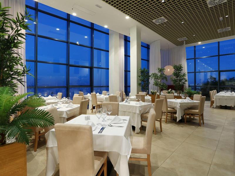 Ruim restaurant met grote ramen, beige stoelen, witte tafelkleden en weelderige groene planten.