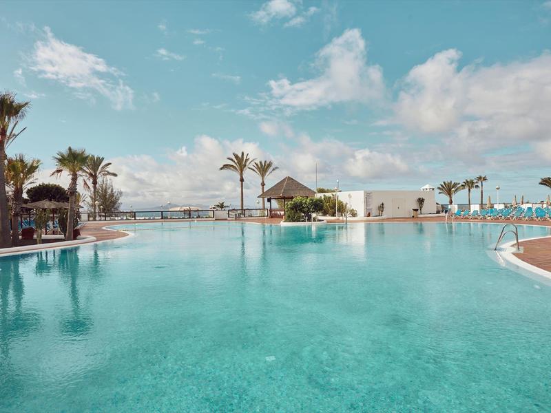 Helder zwembad met ligstoelen en palmbomen onder een blauwe lucht.