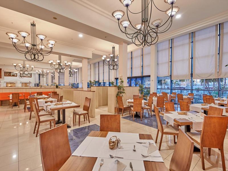 Lichte hotelrestaurant met houten stoelen, witte tafels, kroonluchters en grote ramen.