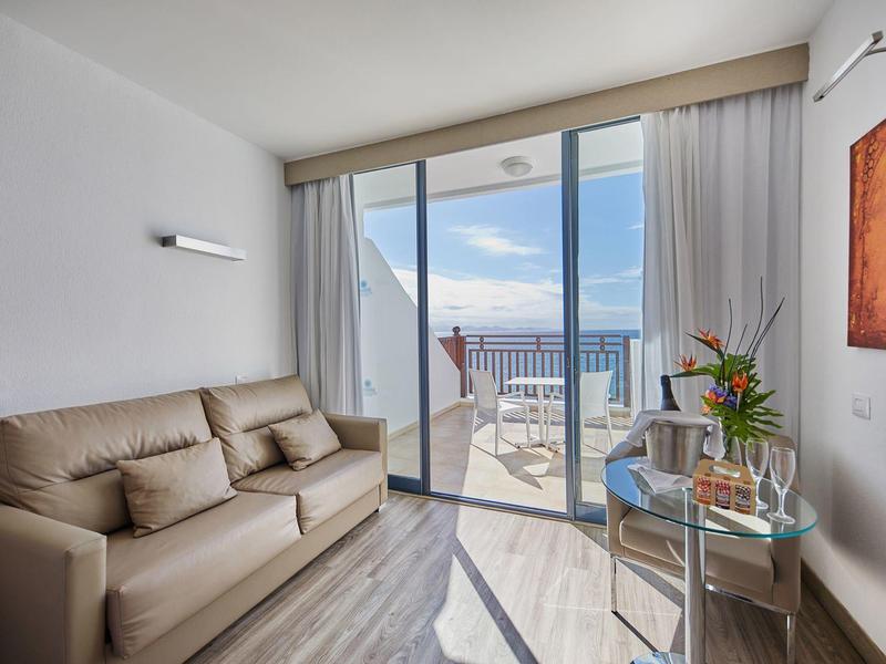 Moderne hotelkamer met bank, glazen tafel en balkon met zeezicht onder een zonnige hemel.