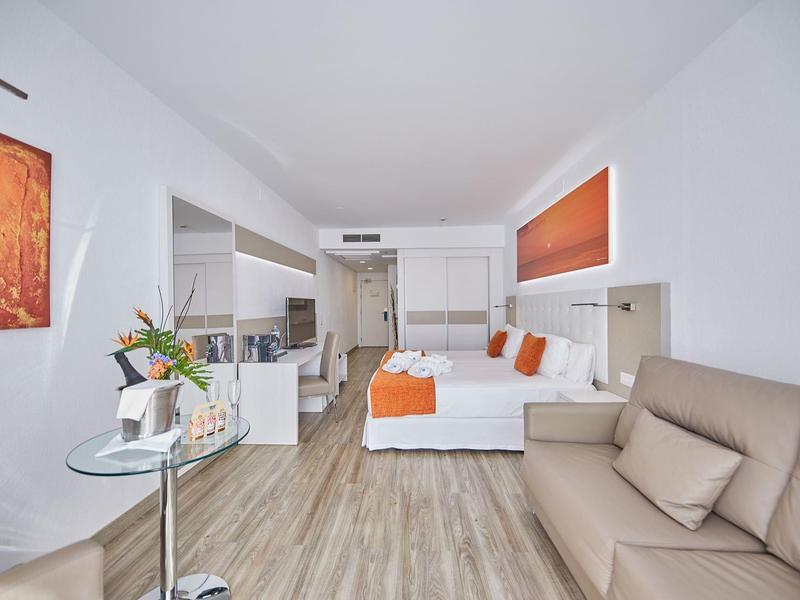 Lichte hotelkamer met kingsize bed, beige bank, kleine tafel en modern decor.