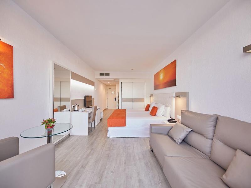 Lichte, moderne hotelkamer met witte muren, oranje accenten, bank, bed en klein tafeltje met bloemen.