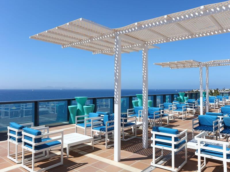 Buitenterras met wit-blauwe zitplaatsen en uitzicht op de oceaan onder pergola's op een zonnige dag.