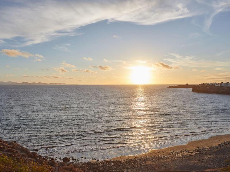 Zon die ondergaat boven een rustige zee met een zandstrand en rotsachtige kustlijn.