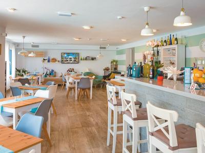 Helles Café mit Holzboden, weißen Stühlen, Tischen und einer Theke mit Getränken und Obstschalen.