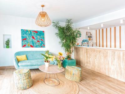 Helles Wohnzimmer mit blauem Sofa, Pflanzen, buntem Wandbild, Holztisch und geflochtenen Poufs.