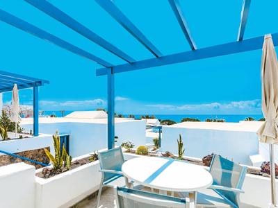 Terrasse mit weißen Möbeln, blauen Kissen und Pergola, Meer im Hintergrund unter blauem Himmel.