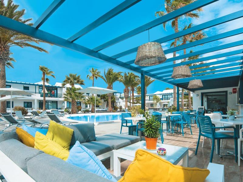 Area relax esterna con divani colorati e tavoli sotto una pergola azzurra vicino alla piscina.