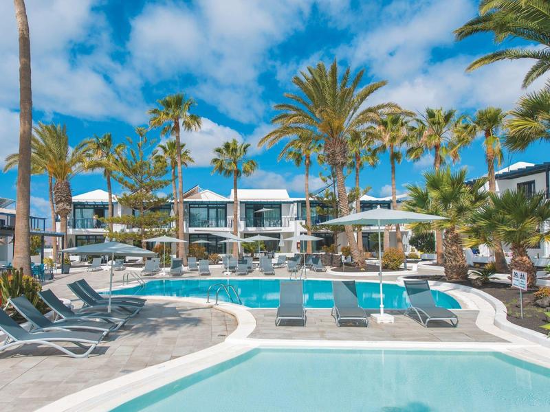 Area piscina di lusso con ombrelloni e lettini, circondata da palme e edifici dell'hotel.