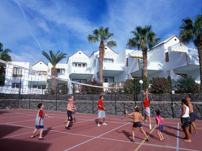 Kinder spielen Basketball auf einem roten Sportplatz vor weißen Häusern und Palmen unter blauem Himmel.