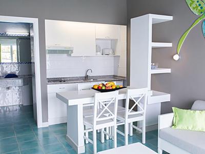 Moderne Küche mit weißer Theke, zwei Stühlen und grauen Wänden, daneben Sitzbereich mit grünem Kissen.