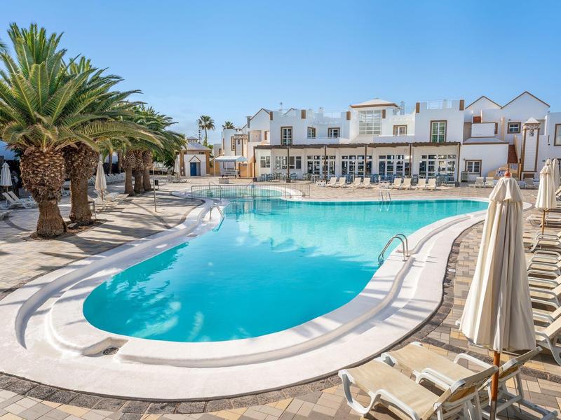 Piscina invitante circondata da palme con un hotel bianco su più piani sullo sfondo.