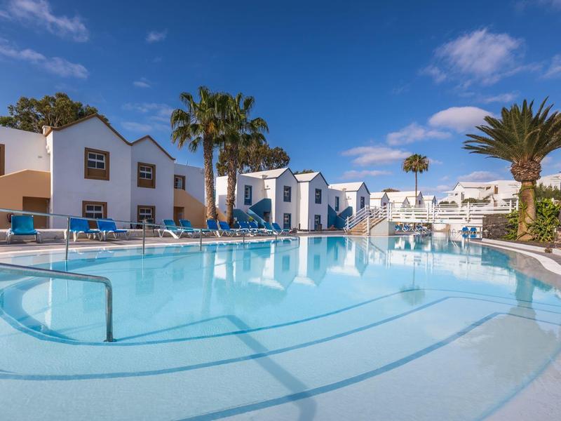 Grande piscina all'aperto con lettini e case vacanze bianche sotto un cielo blu.
