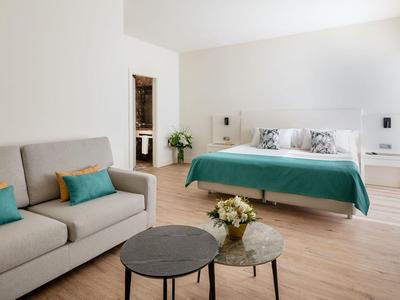 Camera d'hotel luminosa con letto matrimoniale, divano e due tavolini tondi su pavimento in legno.