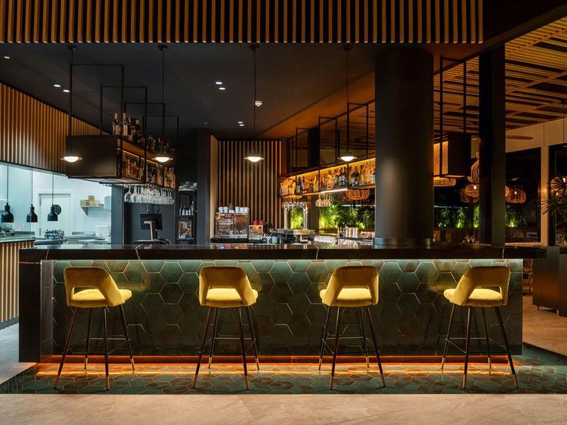 Bar moderna con quattro sgabelli illuminati e cucina aperta sullo sfondo.