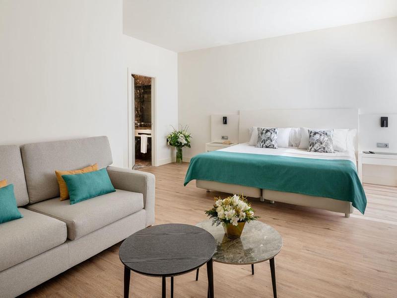 Camera d'hotel luminosa con letto matrimoniale, divano e due tavolini tondi su pavimento in legno.