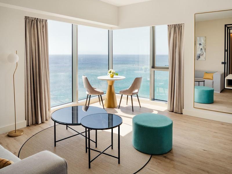 Camera d'hotel moderna con finestre in vetro con vista sul mare, piccolo tavolo con sedie e sgabello.