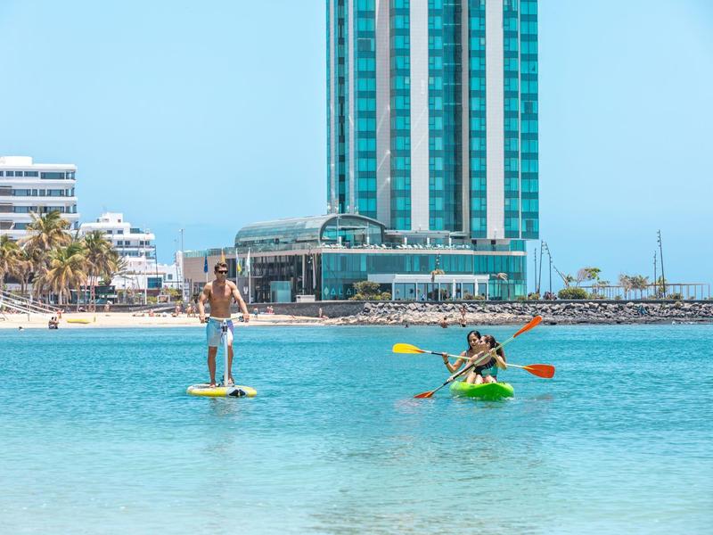 Persone che fanno kayak e paddleboard in acqua limpida davanti a un hotel sulla spiaggia.