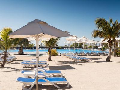 Strand mit weißem Sand, blauen Liegestühlen, Sonnenschirmen und Palmen unter klarem blauen Himmel.