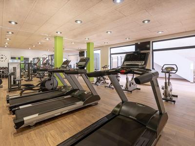 Heller Fitnessraum mit Holzfußboden, Laufbändern, Trainingsgeräten und großen Fenstern.