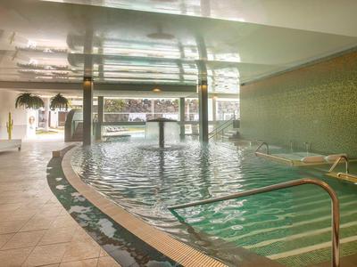 Helles Hallenbad mit runden Becken, Blumenmusterfliesen und Edelstahl-Handläufen am Treppeneingang.