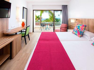 Helles Hotelzimmer mit Doppelbett, roten Akzenten, Holzmöbeln, Balkon und Meerblick.