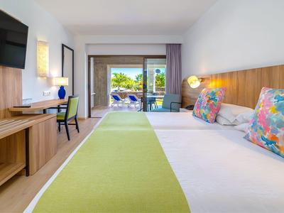 Hell eingerichtetes Hotelzimmer mit großem Bett, grünem Läufer und Balkon mit Meerblick.