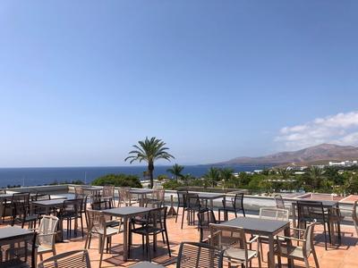 Leere Terrasse mit Tischen und Stühlen, Palmen und Meerblick bei klarem Himmel.