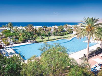 Großer Swimmingpool mit Palmen und Liegestühlen, dahinter Meer und blauer Himmel.