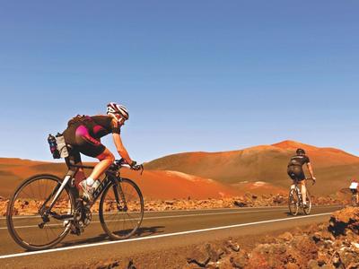 Drei Radfahrer fahren auf einer Straße durch eine rote Wüstenlandschaft unter klarem Himmel.