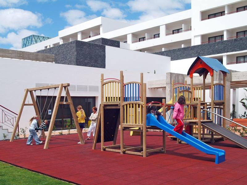 Area giochi per bambini con scivoli, altalene e strutture da arrampicata davanti a un hotel moderno.