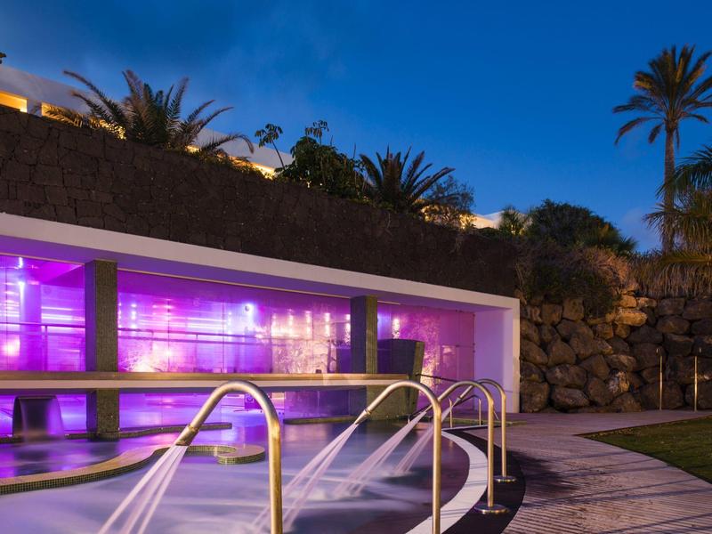 Piscina illuminata con corrimano e palme al crepuscolo in un giardino d'hotel.
