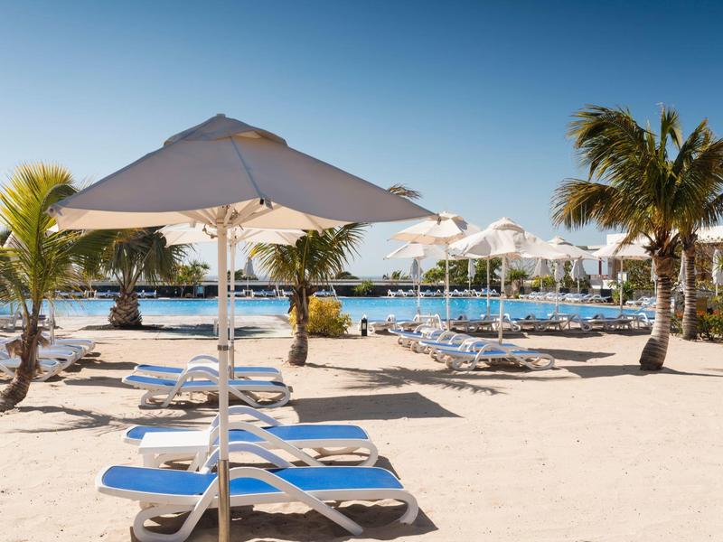 Area spiaggia con lettini, ombrelloni e palme sotto un cielo limpido.