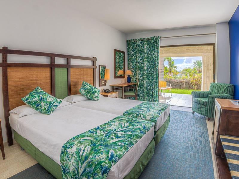 Camera d'albergo luminosa con due letti, biancheria verde e accesso a una terrazza con vista.