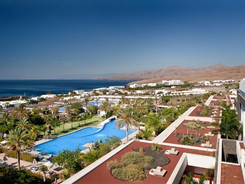 Vista di un complesso alberghiero con piscina, palme e mare sullo sfondo con cielo sereno.