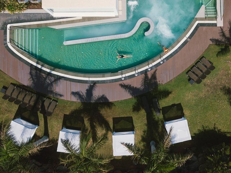 Veduta aerea di una piscina d'hotel curva con lettini e capanne di paglia sul prato.