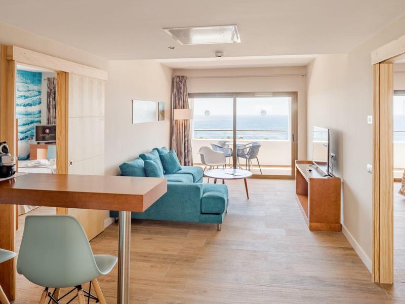 Soggiorno luminoso con divano blu, pavimento in legno e vista sul mare da grandi finestre.