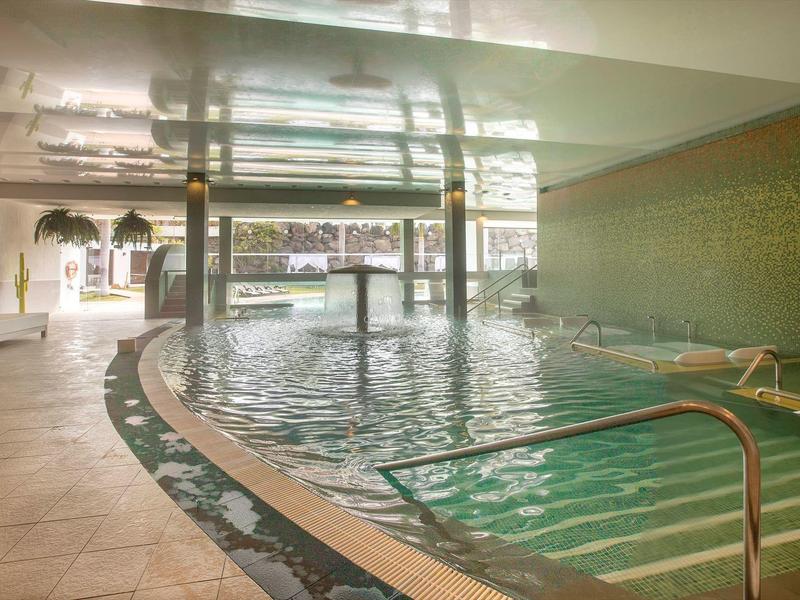 Interno di una piscina illuminata con scale e area relax accanto a una parete di vetro.