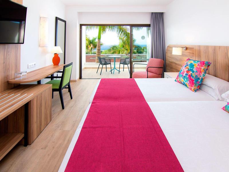 Camera d'hotel luminosa con due letti, coperta rossa e terrazza con vista su palme.